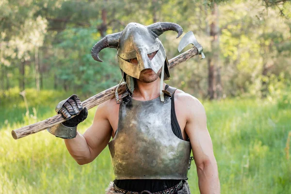 stock image Portrait of a warrior in a horned helmet, a steel breastplate with a two-handed ax in his hands, posing against the backdrop of the forest. 