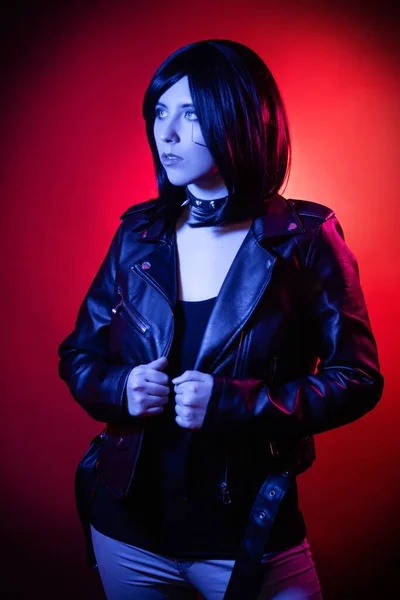 Portrait of young woman with bob haircut in black leather jacket illuminated with blue light posing isolated on dark background with red illumination, cyberpunk concept.