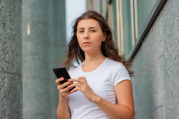 一个穿着白色T恤 手里拿着智能手机的年轻女子的画像 背对着墙和柱子摆姿势 模仿者 — 图库照片