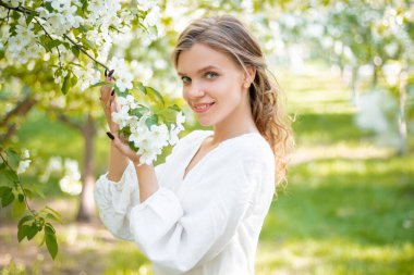 Beyaz elbiseli genç bir kadının portresi elinde çiçeklerle çiçek açan elma ağaçlarının arka planında. Kız, bahar parkında çiçeklerin arka planında poz veriyor..
