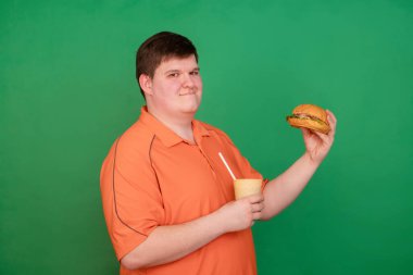 Büyük bir hamburger yiyen şişman bir adamın portresi ve yeşil arka planda izole edilmiş bir kamışla bir bardaktan soda içmesi. Krom anahtar, yeşil ekran. Fast food ve obezite kavramı.