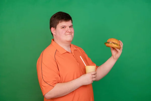 Portrait of a fat guy eating a big hamburger and drinking soda from a glass with a straw, isolated on a green background. Chroma key, green screen. The concept of fast food and obesity.