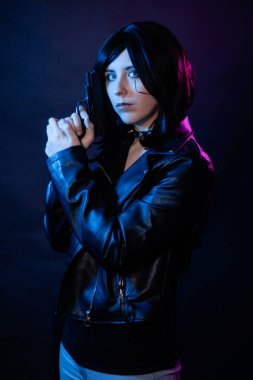 Siyah deri ceketli, çivili yakalı, elinde silahla siyah bir arka planda izole edilmiş, siber punk konsepti olan genç bir kadının portresi..