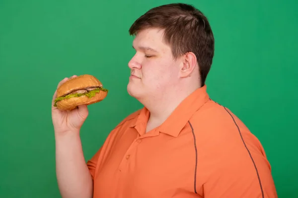 Portrait of a fat guy with a big hamburger in his hands, isolated on a green background. Chroma key, green screen. Fast food and obesity concept.