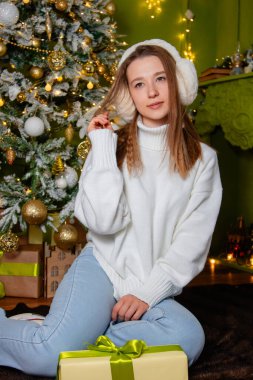 Beyaz kazak giymiş, kulaklık takmış ve kot pantolon giymiş genç bir kadın elinde bir Noel ağacının yanında oturuyor..