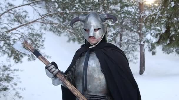 一个戴着角形头盔的中世纪幻想战士的画像 钢制的胸牌 双手拿着一把双手斧子的链子 在冬季森林的背景下摆姿势 照相机运动 — 图库视频影像