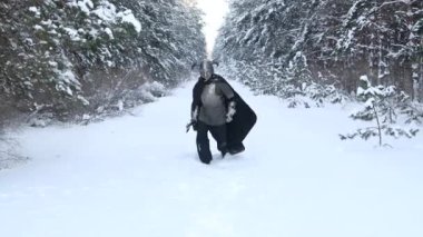 Boynuzlu miğferli bir ortaçağ fantezi savaşçısı, çelik bir göğüs zırhı ve elinde iki elli bir baltayla kış ormanına saldırmak için koşuyor. Ses ile video, seçici odak.