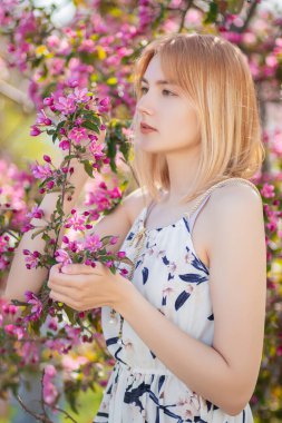 Elbise giymiş çekici bir genç kadının portresi çiçek açan pembe elma çiçeklerine karşı poz veriyor..