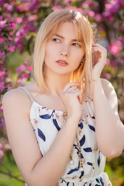 Elbise giymiş çekici bir genç kadının portresi çiçek açan pembe elma çiçeklerine karşı poz veriyor..