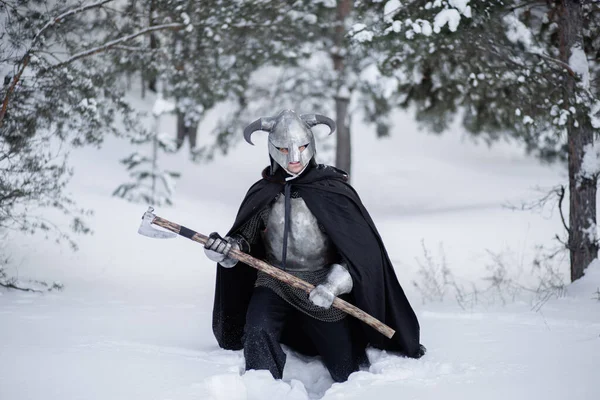 A medieval fantasy warrior in a horned helmet, a steel breastplate and chain mail with a two-handed ax in his hands, walks through snowdrifts in a winter forest.