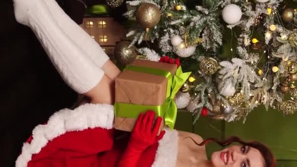 一个穿着红色连衣裙 手里拿着礼物的年轻女子坐在圣诞树下的画像 女式圣诞老人套装垂直视频 相机运动 选择性聚焦 — 图库视频影像