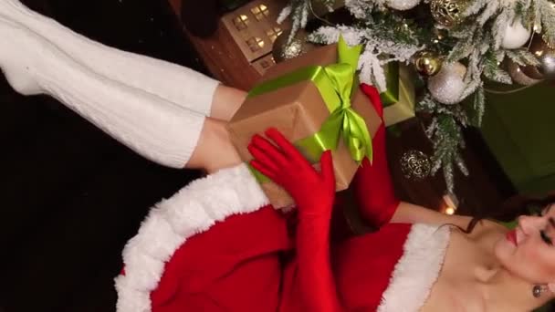 一个穿着红色连衣裙 手里拿着礼物的年轻女子坐在圣诞树下的画像 女式圣诞老人套装垂直视频 相机运动 选择性聚焦 — 图库视频影像