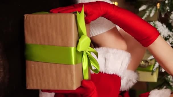 女性手拿着礼物的特写 一个穿着圣诞圣塔服装的女人靠着圣诞树的背影 垂直视频 相机运动 选择性聚焦 — 图库视频影像