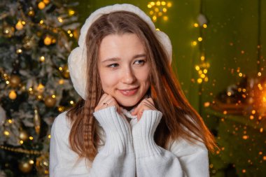 Beyaz süveter ve kalın kulaklıklar takmış, altın bir bokeh ve bir Noel ağacına poz veren genç bir kadının yakın plan portresi.