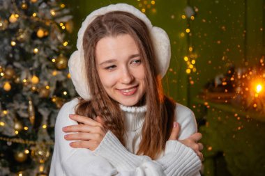 Beyaz süveter ve kalın kulaklıklar takmış, altın bir bokeh ve bir Noel ağacına poz veren genç bir kadının yakın plan portresi.
