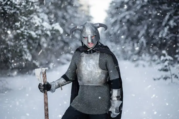 stock image Portrait of a medieval fantasy warrior in a horned helmet, steel cuirass, chain mail with a two-handed ax in his hands, posing against the backdrop of a winter forest.