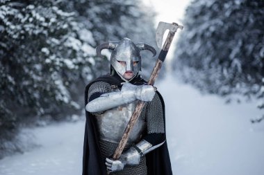 Portrait of a medieval fantasy warrior in a horned helmet, steel cuirass, chain mail with a two-handed ax in his hands, standing in a fighting position against the backdrop of a winter forest. clipart