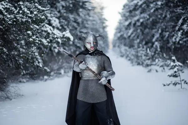 stock image Portrait of a medieval fantasy warrior in a horned helmet, steel cuirass, chain mail with a two-handed ax in his hands, standing in a fighting position against the backdrop of a winter forest.