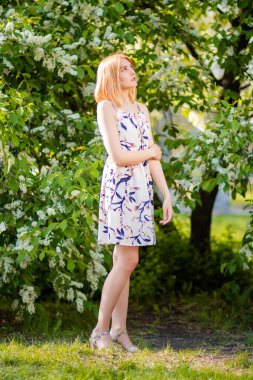 Elbise giymiş, beyaz elma çiçeklerinin arka planında poz veren çekici bir genç kadının tam boy portresi..