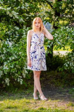 Elbise giymiş, beyaz elma çiçeklerinin arka planında poz veren çekici bir genç kadının tam boy portresi..