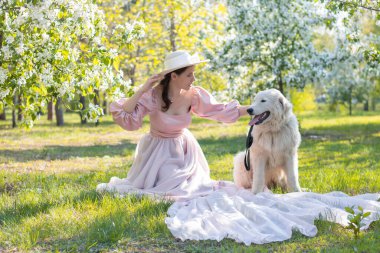 Pembe elbiseli, tren ve şapkalı genç bir kadının portresi, büyük beyaz tüylü bir köpeğin yanında bir bahar parkının arka planında oturuyor ve çiçek açıyor..