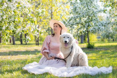 Pembe elbiseli, tren ve şapkalı genç bir kadının portresi, büyük beyaz tüylü bir köpeğin yanında bir bahar parkının arka planında oturuyor ve çiçek açıyor..