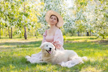 Portrait of a young woman dressed in a pink dress with a train and a hat, sitting next to a large white fluffy dog against the backdrop of a spring park and blooming flowers. clipart