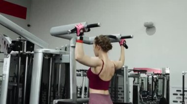 Spor salonundaki bir makinede barfiks çeken genç atletik kadın. Pürüzsüz kamera hareketi, seçici odak.