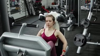 Genç, atletik bir kadın spor salonundaki bir makineye bacak presi yapıyor, bacaklarını çalıştırmak için. Kamera hareketi, seçici odak.