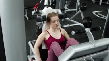 Genç, atletik bir kadın spor salonundaki bir makineye bacak presi yapıyor, bacaklarını çalıştırmak için. Kamera hareketi, seçici odak.