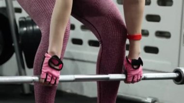 Genç bir atletik kadın spor salonunda halterle ağırlık kaldırma, sırt ve bacak antrenmanı yapıyor. Kamera hareketleri, seçici odaklanma..
