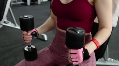 Spor salonunda elinde halterlerle bir bankta otururken egzersiz malzemelerinin arka planında poz veren genç bir kadın. Kamera hareketi, seçici odak.