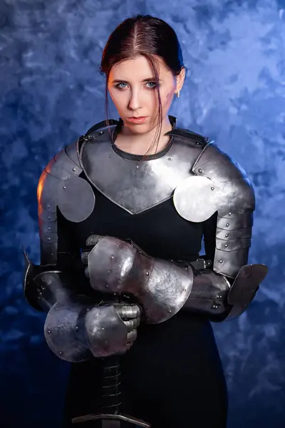 stock image Portrait of a young woman with scars on her face, dressed in black clothes with steel knightly hands and a gorget, holding a bastard sword, posing against an abstract background. Medieval fantasy girl