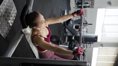 Dikey video, spor salonundaki bir bankta halterle ağırlık kaldıran genç atletik kadın. Seçici odak yumuşak hareket.