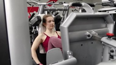 Genç, atletik bir kadın spor salonundaki bir makineye bacak presi yapıyor, bacaklarını çalıştırmak için. Kamera hareketi, seçici odak.