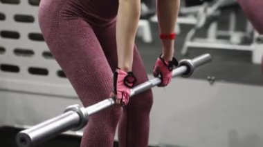 Genç bir atletik kadın spor salonunda halterle ağırlık kaldırma, sırt ve bacak antrenmanı yapıyor. Kamera hareketleri, seçici odaklanma..
