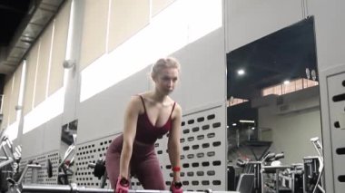 Genç bir atletik kadın spor salonunda halterle ağırlık kaldırma, sırt ve bacak antrenmanı yapıyor. Kamera hareketleri, seçici odaklanma..