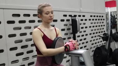 Genç bir atletik kadın spor salonunda egzersiz malzemelerinin arka planında otururken poz veriyor. Kamera hareketi, seçici odak.