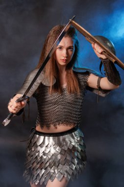 Elinde kılıç ve kalkanla karanlık bir arka planda izole edilmiş mavi bir ışık ve duman içinde duran ortaçağ kadın savaşçısının portresi..