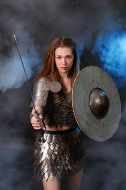 Elinde kılıç ve kalkanla karanlık bir arka planda izole edilmiş mavi bir ışık ve duman içinde duran ortaçağ kadın savaşçısının portresi..