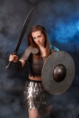 Elinde kılıç ve kalkanla karanlık bir arka planda izole edilmiş mavi bir ışık ve duman içinde duran ortaçağ kadın savaşçısının portresi..