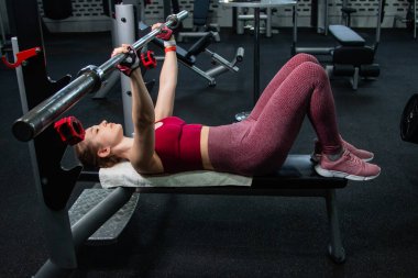 Spor salonundaki bir bankta halterle ağırlık kaldıran genç atletik kadın..