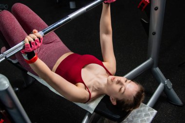 Spor salonundaki bir bankta halterle ağırlık kaldıran genç atletik kadın..