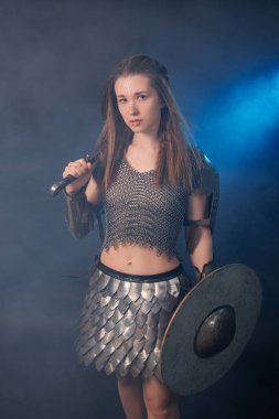 Elinde kılıç ve kalkanla karanlık bir arka planda izole edilmiş mavi bir ışık ve duman içinde duran ortaçağ kadın savaşçısının portresi..