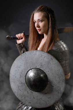 Elinde kılıç ve miğfer olan, karanlık bir arka planda dumanın içinde duran, ortaçağ kadın savaşçısının portresi..