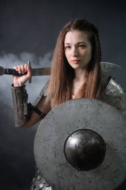 Elinde kılıç ve miğfer olan, karanlık bir arka planda dumanın içinde duran, ortaçağ kadın savaşçısının portresi..