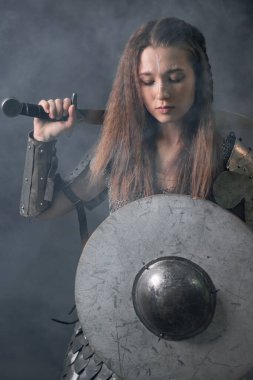 Elinde kılıç ve miğfer olan, karanlık bir arka planda dumanın içinde duran, ortaçağ kadın savaşçısının portresi..