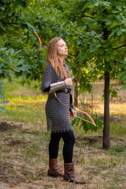 Ortaçağ savaşçısı kadın, zırhlı ve elinde yayıyla yaz ormanı zemininde..