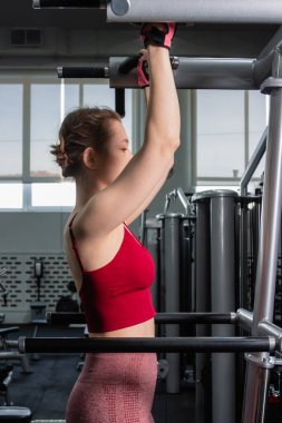 Spor salonundaki bir makinede barfiks çeken genç atletik kadın. Kol ve sırt egzersizi.