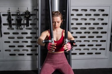 Genç atletik güzel bir kadın tam teçhizatlı bir spor salonunda göğüs makinesinde egzersiz yapıyor..